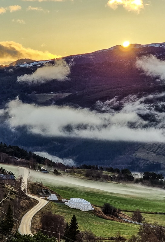 Landskapsbilde av Vågå med soloppgang, fjell og lett morgentåke over jordene.